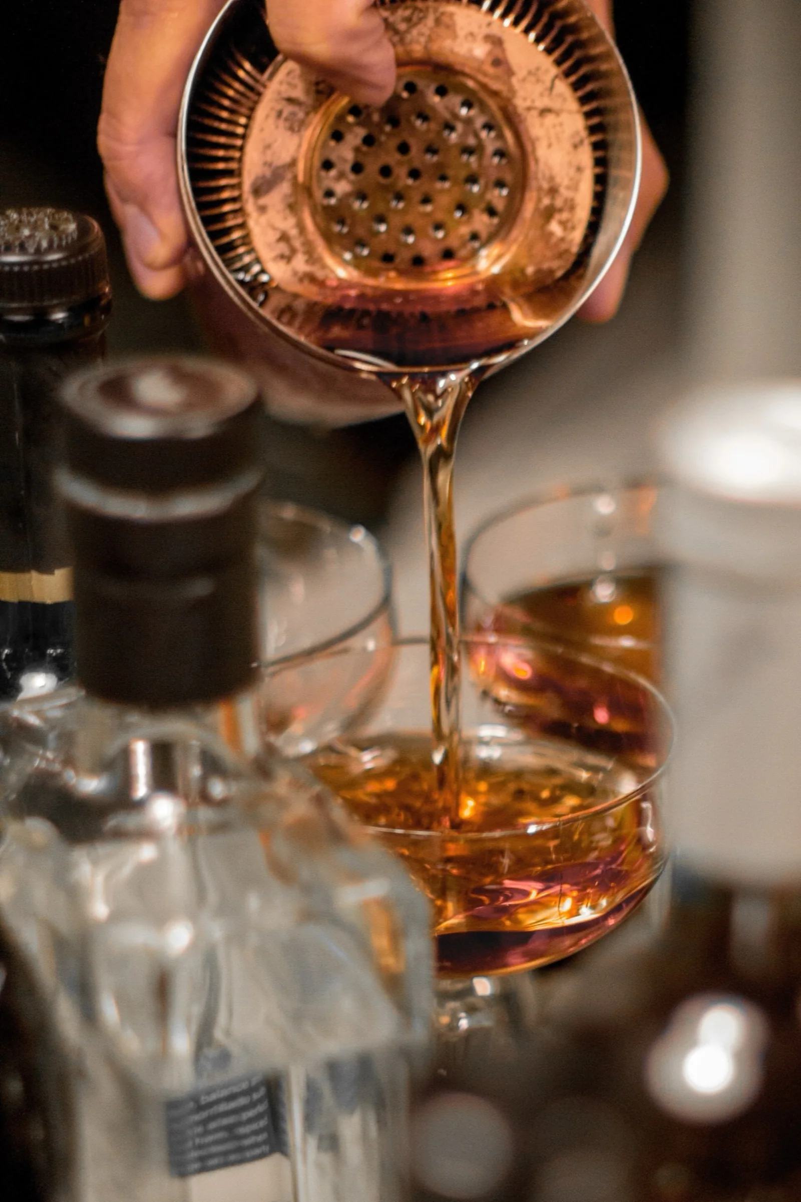 Bartender straining a stirred amber cocktail through a strainer into a coupe glass